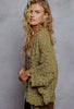 A woman with long wavy blonde hair models a textured, olive green crochet cardigan with scalloped edges, posed against a neutral gray studio backdrop.