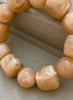 A close-up of a beaded bracelet made of glossy, irregular peach-beige stone or shell-like beads resting on a textured beige fabric surface.