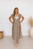 A woman modeling a short-sleeve leopard-print maxi dress with a V-neckline and white lace detailing on the bodice, standing in a bright room with a white paneled wall, light wood floor, a woven basket, and a vase with dried grass.