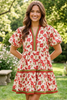 A woman wearing a short puff-sleeve tiered dress with a red poppy floral print on a cream background, standing in a sunny garden setting.