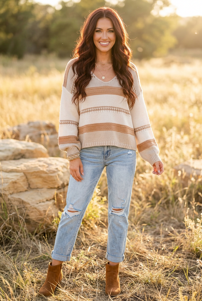 A woman stands outdoors in a grassy field at sunset wearing a cream and tan striped knit sweater, light blue ripped jeans, and brown ankle boots.