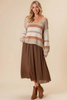 A woman modeling a cozy knit sweater with horizontal stripes in cream and tan, paired with a brown midi skirt and brown ankle boots, against a solid beige studio background.