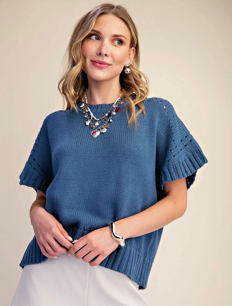 A woman wearing a blue short-sleeve knit sweater with textured eyelet details on the sleeves, styled with white pants and statement jewelry against a plain beige background.