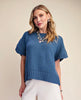 A woman modeling a blue short-sleeve knit top with textured eyelet sleeves and a ribbed hem, paired with white pants, against a plain beige studio background.