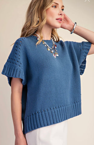 A woman wearing a blue short-sleeve knit sweater with textured openwork details on the sleeves and a ribbed hem, paired with white pants and layered necklaces, against a plain beige background.