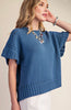 A woman wearing a blue short-sleeve knit sweater with textured openwork details on the sleeves and a ribbed hem, paired with white pants and layered necklaces, against a plain beige background.