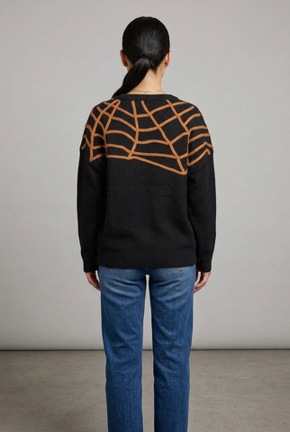 A woman viewed from behind wearing a black knit sweater with a large orange spiderweb pattern across the shoulders and upper back, paired with blue jeans, against a neutral studio background.