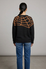 A woman viewed from behind wearing a black knit sweater with a large orange spiderweb pattern across the shoulders and upper back, paired with blue jeans, against a neutral studio background.