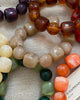 A close-up of multiple beaded bracelets arranged on a textured beige fabric background. The bracelets feature chunky, polished beads in various colors including amber brown, cream, orange, green, dark green, and purple.