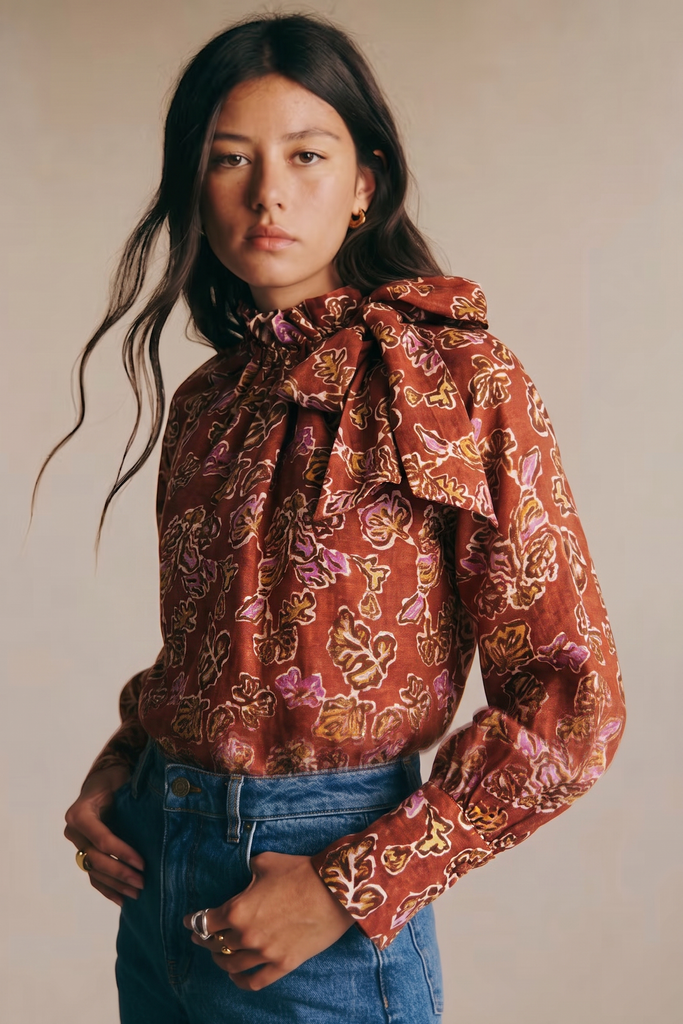 A woman wearing a rust-orange long-sleeve blouse with a large ruffled bow at the neck and an allover floral pattern in pink, purple, and gold, paired with blue jeans against a plain beige background.
